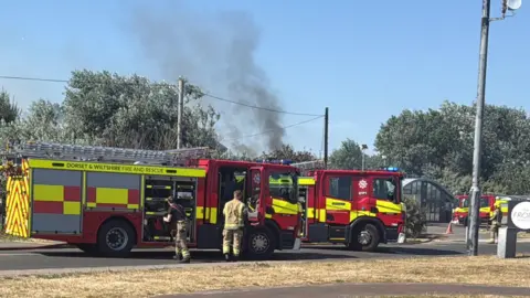 Fire engines parked on a road with smoke rising in the background