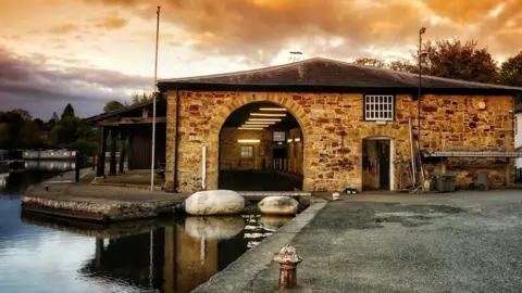 Ian Roberts A stone building with a large arched entrance and a wooden canopy to the left next to the canal.