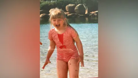 Helen Stroud A young girl wearing a one-piece red and white striped swimming costume stands in the sea, exclaiming happily. She has long blonde hair and a fringe, in a ponytail. You can see a collection of different sized moles across her legs.