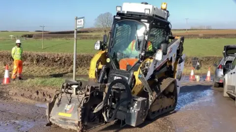 A driveable machine with caterpillar tracks and a device on the front to help with pothole repair. It is in use on a road in a rural area with a field and blue sky behind.