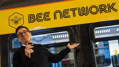 TFGM Andy Burnham, a man with short dark hair and glasses, stands with a microphone in front of a train which has a yellow sign which reads Bee Network.