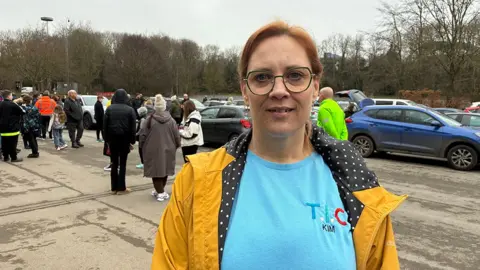 A woman with short red hair and wearing glasses is standing in a car park with people congregating behind her. She is wearing a blue t-shirt with a logo that spells TIC underneath an open yellow coat.