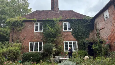 Baschea Walsh stands holding a mug in front of a cottage. It has two floors and four windows, parts of the front of the building are covered in ivy. To the right of Baschea, there is a life-sized model of a sheep. 