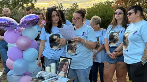 George King/BBC A group of women wearing blue T-shirts with Courtney Mitchell's face on them. They are standing next to some balloons. One of the women, in the centre of the image, is holding a microphone and reading from a piece of paper. 
