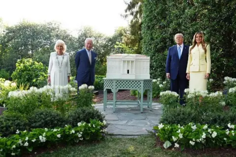 PA King Charles III and Queen Camilla join US President Donald Trump and First Lady Melania, all dressed formally, in a green garden, beside a beehive in the shape of the White House