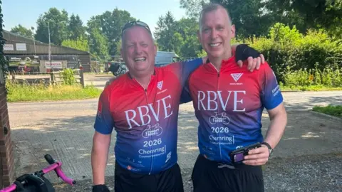 RBVE Two men in a red and blue cycling gear. There is a bicycle next to them.