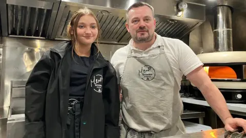 Noel O'Hagan and Ellie Hill standing inside their mobile pizza business. A pizza oven can be seen behind Noel as well as shiny kitchen surfaces all around. Ellie has short brown hair and is wearing a black jacket, black T-shirt and black apron. Noel has short grey hair and a short grey beard and he is wearing a white T-shirt and grey apron.