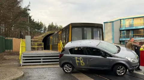 A grey and yellow Metro train carriage is half in a tunnel, half out. A grey car is pushed on its side in front of it. There are two firefighters in the far right corner, one of them is pointing at something out of shot.