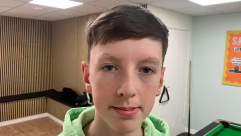 A teenage boy, pictured from the shoulders up, with short brown hair. He is wearing a bright green hoodie. 