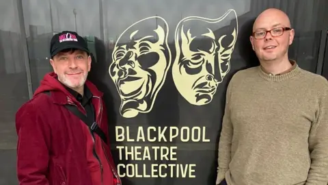 Blackpool Theatre Collective Two men stand outside a glass window which has a sign of two theatre masks, with writing underneath which reads 'Blackpool Theatre Collective'