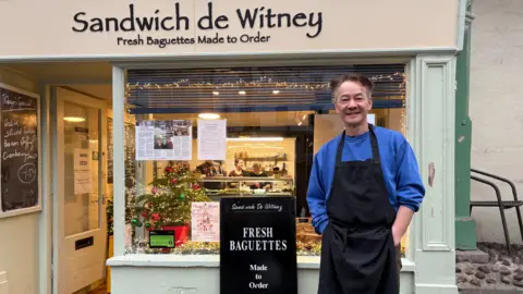 The owner of a sandwich shop stands in front of his shop window. Henry Mo wears a blue sweater and black apron. In his window is a small Christmas tree and warm coloured fairy lights. In the back of the shop you can spot the employee's behind the counter.