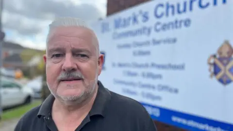 Helen Mulroy/BBC Councillor and vicar Charles Royden, standing outside a sign for St Mark's Church, Bedford. He has grey hair, a bear and is wearing a dark polo style T-shirt. The sign is blurred behind him. 