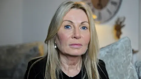 Janet Taylor is wearing pearl earrings and a black T-shirt and sitting on a grey couch in her living room. She is looking straight at the camera. 