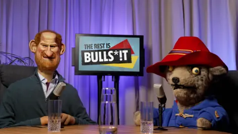 Avalon Puppets of Prince Harry and Paddington hosting a podcast. They are sat at a table in a studio with a TV screen behind them.