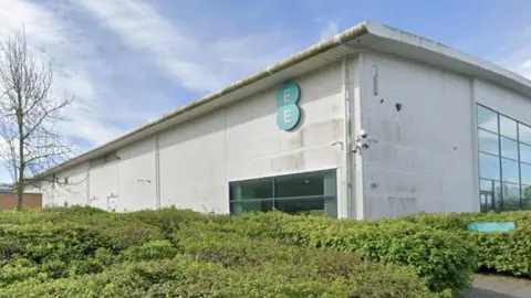 EE call centre at Doxford Park in Sunderland. It is a white facility that has green moss growing on its side with a blue EE logo fixed on one of its faces. There are several security cameras attached to the corner of the building. There are large green bushes at the front of building.