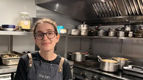 Ella Hardy in the silver chrome kitchen of The Yan At Broadrayne in Grasmere. She has dark hair tied back and dark eyes. She wears round glasses with a dark frame, a black top and a denim apron with leather straps. She is smiling at the camera. Behind her is a cooker with some pots on the stove and others stored on a shelf above.