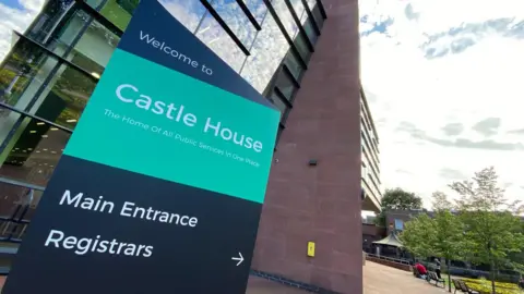 A green and black totem, with white writing that says Welcome to Castle House. There is a large building behind it, with a band stand and benches in the distance, as well as trees.