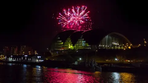 Purple, red and blue fireworks explode over the Sage Gateshead, a large glass building alongside the River Tyne.