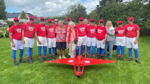 Barlow Red Barrows A row of people stood arm in arm wearing red tops, white shorts and blue bottoms on their lower legs. A red wheelbarrow with plane wings is in front of them.