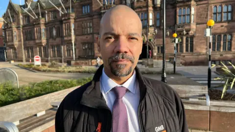 Delcan Downes stands outside the headquarters of Coventry City Council, an ornate sandstone building with lead lined windows. Delcan wears a black jacket featuring the GMB logo and a white shirt with pink tie.