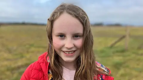 Young girl smiling at the camera. She has long fair hair and is wearing a red coat