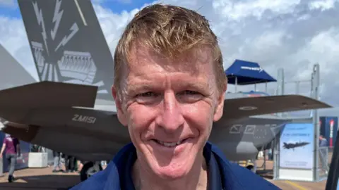 A head and shoulders picture of Tim Peake smiling into the camera while outside on a sunny day. He has short ginger hair and is wearing navy overalls. In the background is a military jet.