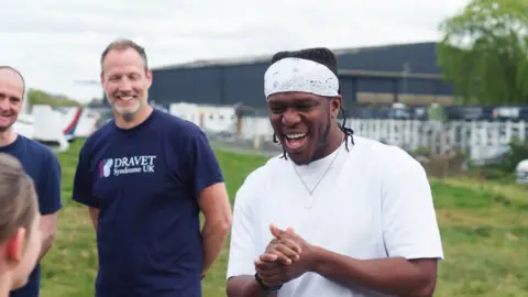 A man, KSI amiles as he stands in a field with his hands together, he wears a white tshirt and bandana.