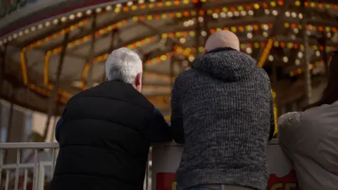Two men with their backs to the camera look at a funfair carousel