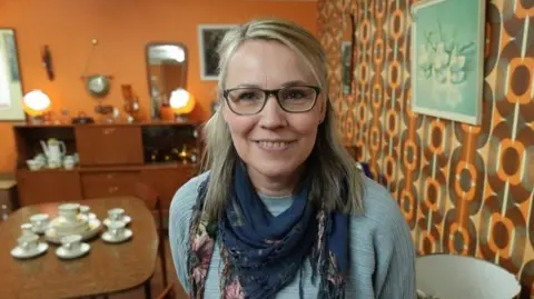 A woman with blonde hair and glasses is standing in a shop with mid-century wallpaper and furniture 
