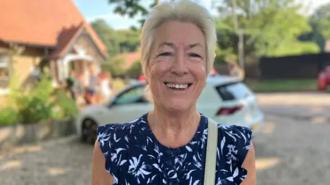 Owen Sennitt/BBC A woman smiles towards the camera on a sunny day wearing a blue flowery top outside a community centre