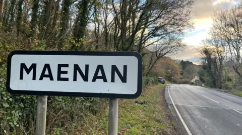 BBC Road sign for the village of Maena, with a country road surrounded by trees