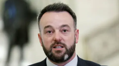 Reuters Colum Eastwood has short dark hair with white sides, and a dark beard with two white patches at the front. He is wearing a grey suit jacket, a white shirt and a dark blue with white polka dots tie.