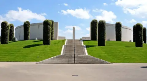Getty Images Neat and tidy grass and cropped trees surround a long, sweeping concrete pathway up to a memorial at the site. There is a circular design to it with a large monument or plinth in the middle.
