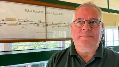 Andrew Turner/BBC Steven Ashling standed in front of a Reedham junction railway line sign and is wearing circular glasses and a green polo shirt.