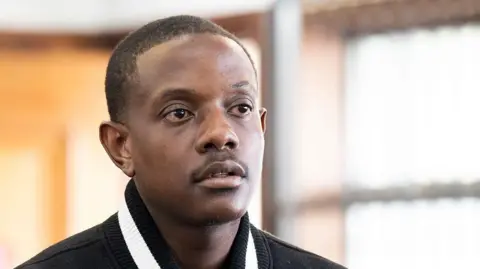Close-up of Bellarmine Mugabe. The partial black and white collar and black shoulders of his jacket can be seen. His is standing indoors.