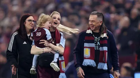 Getty Images A group of four people, on the left is a woman with dark red hair and glasses, wearing a black tracksuit. In the centre is a  woman with blonde hair wearing an Aston Villa scarf, holding her toddler and smiling widely, and to her right is a man with long hair who is looking at the toddler and smiling, he is also wearing an Aston Villa scarf. 