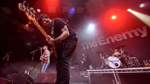 A man with brown hair is bent over with a guitar in his arms. He is grimacing as he plays, and has tattoos on his arm. Behind him to his right is a man with a guitar singing into a microphone on a stand. Behind them, there is a man with a guitar and next to him is a man playing the drums. On a black backdrop there is white writing that says "the enemy"