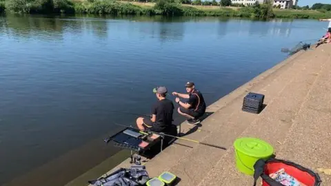 Canal and River Trust Two anglers, surrounded by equipment, sit on a series of concrete steps by a river
