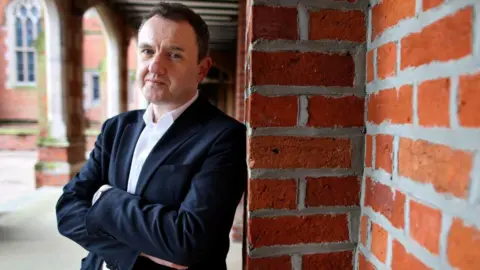 Getty Images A man with a white shirt and a dark suit jacket leans against a red-brick wall. He has his arms crossed and he is looking at the camera. 