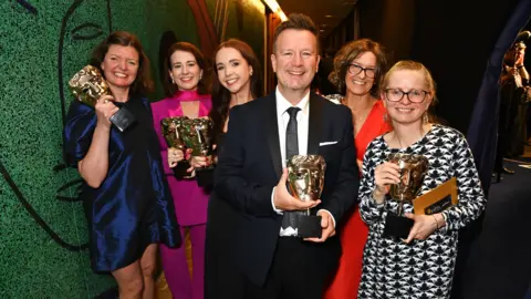 Getty Images A line of eight people wearing formal clothes for an award ceremony. Six of them are holding Bafta statuettes. Alison Howe is far right wearing a black and white dress.