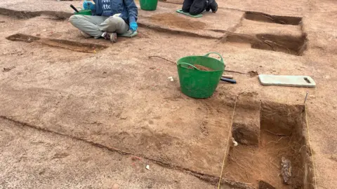 Tony Fisher/BBC Several shallow trenches and test pits are carefully laid out across the excavation area. Clean edges and string lines mark features being recorded and measured. A kneeling archaeologist uses hand tools to precisely uncover and document remains. Buckets and equipment nearby show the methodical process of removing soil layer by layer.