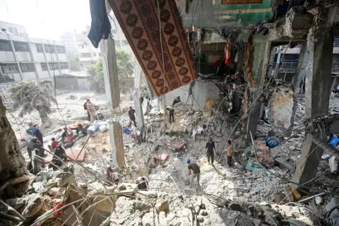 NurPhoto via Getty Images People inspect the damage around a huge crater in a blown-out school caused by Israeli airstrikes, with damage and rubble all around.