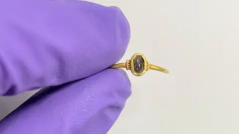 A close up of two fingers wearing a purple glove holding a gold ring. The ring has the remains of a dark red cabochon stone, which sits inside an oval bezel, with an aperture that is decorated with incised circles and grooves on the ring's shoulders. 