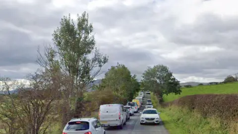 A straight, narrow country road near the proposed caravan park with a long line of traffic on the left hand side and fewer vehicles travelling towards the camera. The road is lined by a few trees and hedges, with wild green fields either side and mountains in the distance.