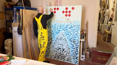 Mr Reynolds is again in his studio, wearing the same outfit. He has his back to the camera and is facing his artwork, called 'Jaws' which is a braille imitiation of the original movie poster. Mr Reynolds is holding his hand up to the top left corner of the artwork to read the braille. The poster shows red dots, which spell out Jaws in braille. The outline of a swimmer in the water and the head of a shark emerging from the bottom of the poster.