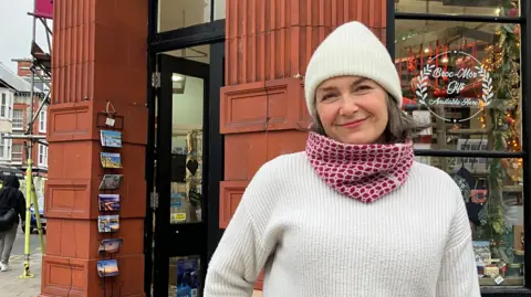 Berith Lochery, wearing a white knit hat and jumper and a red and white scarf, standing outside her shop on Aberystwyth's high street