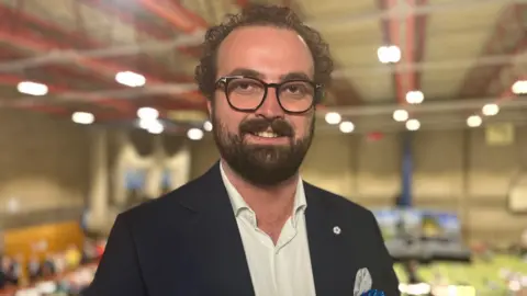 Phil Shepka/BBC Ross Martin stares at the camera. He weats a black jaket with a white shirt. In the background is a sports hall being used for an election count