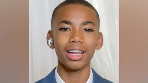 Head shot of Jamal Coombes who is wearing a white shirt beneath a blue jacket. He has a white air pod in one ear