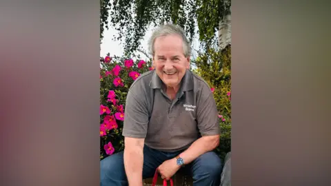 A man smiles at the camera as he sits down in front of a green bush with pink flowers. He has grey hair and is wearing blue jeans and a brown polo shirt with "Wroxham Barns" written on its left-hand side. 