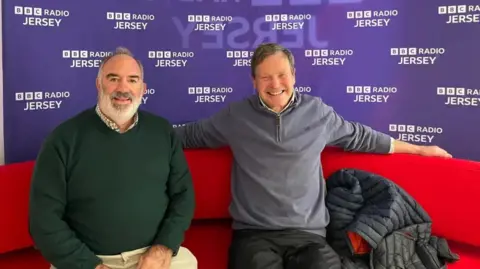 Bob De La Haye and John Howell sit on a red sofa with a purple and white BBC Radio Jersey sign behind them. Mr De La Haye is wearing a chequered shirt with a green jumper. Mr Howell is wearing a blue fleece.